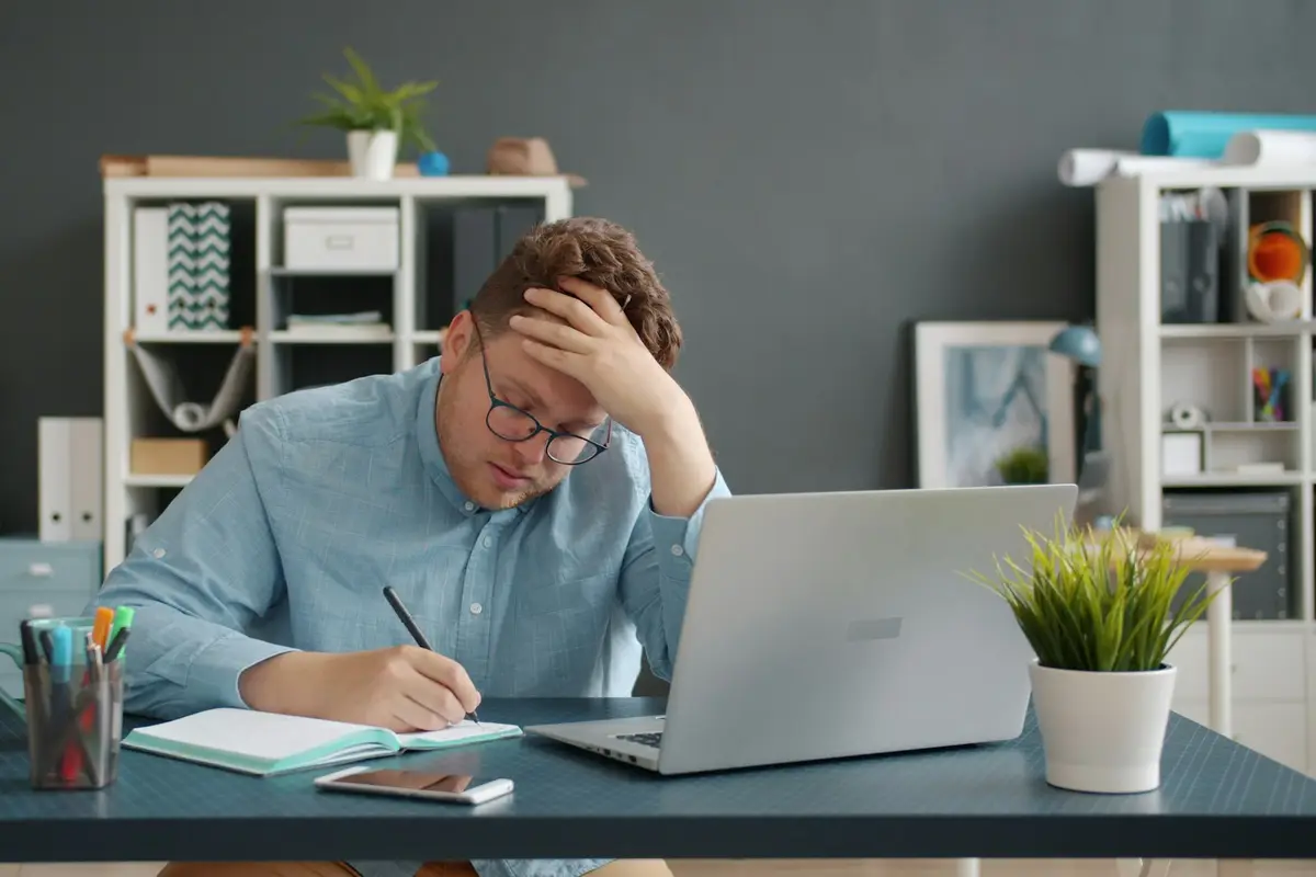 Small business owner at an office desk with a laptop, writing in a notebook and holding his head while working out how to repay an overdue loan